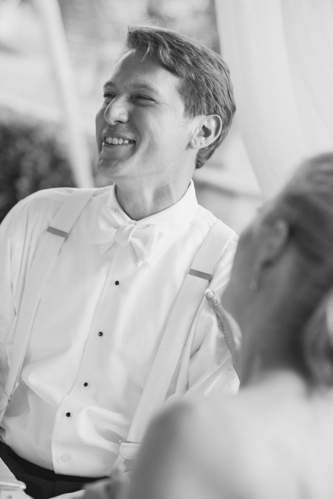 A man in a tuxedo with suspenders and a bow tie smiles warmly, seated next to a woman whose face is out of focus in the foreground. The image is in black and white, capturing a joyful moment.