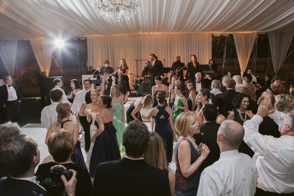 A lively wedding reception scene with guests dancing and socializing under a draped white tent, while a live band performs on stage in the background. The atmosphere is festive and elegant.