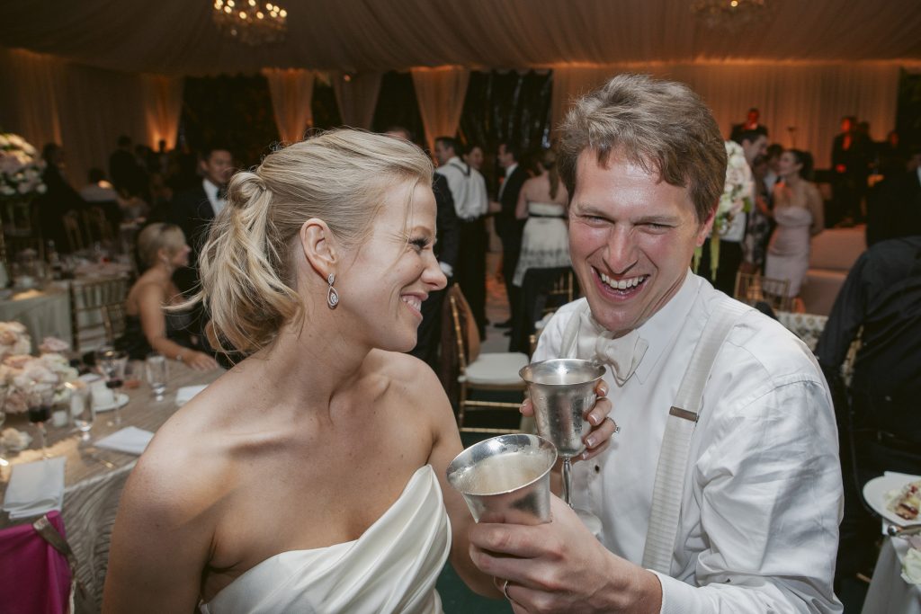 A smiling couple dressed in formal attire laugh and hold metal cups at a lively indoor event, surrounded by other guests seated at decorated tables.