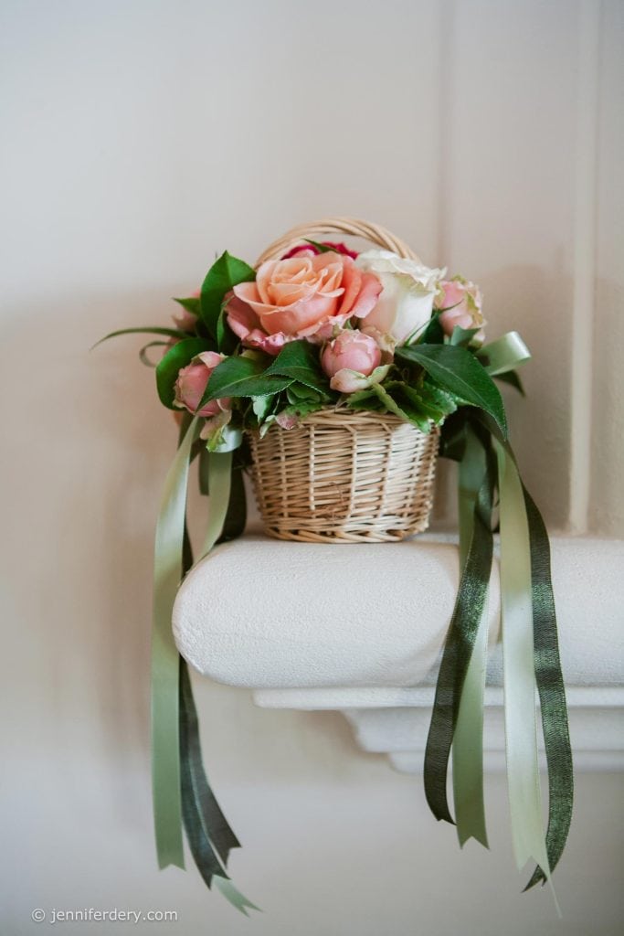 A small wicker basket filled with pink roses, green leaves, and long green ribbons sits on a white ledge against a light-colored wall.