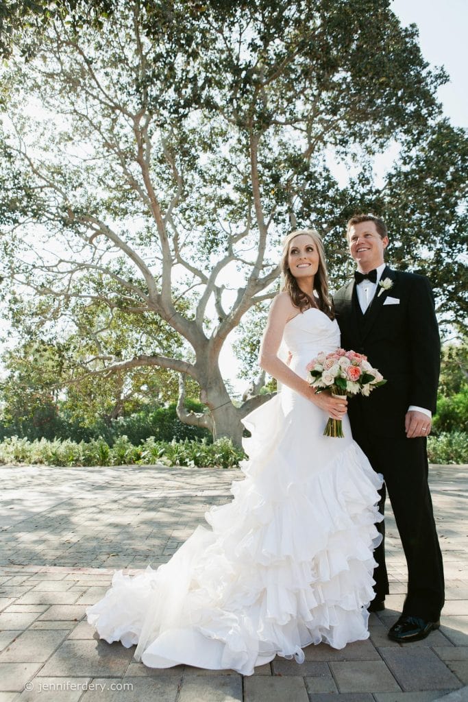 A bride in a white gown and a groom in a black tuxedo stand smiling outdoors on a sunny day in San Diego, with a large tree and greenery in the background. The bride holds a bouquet of pink and white flowers.