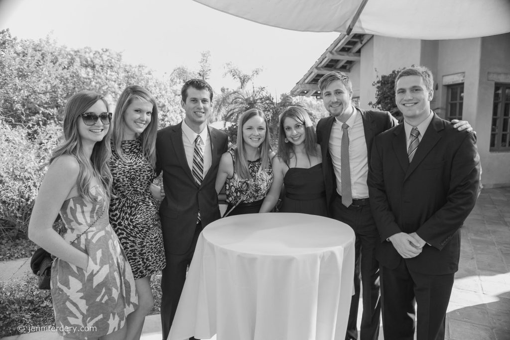 A group of seven young adults dressed in formal attire pose and smile together around a round, white-covered table outdoors on a sunny day.