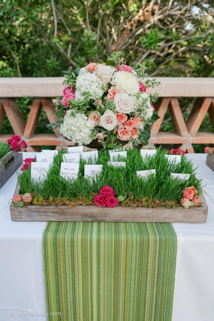 A table decorated with a green striped runner displays trays of grass holding white place cards, accented with pink and peach roses. A large bouquet of white and pink flowers sits in the center.