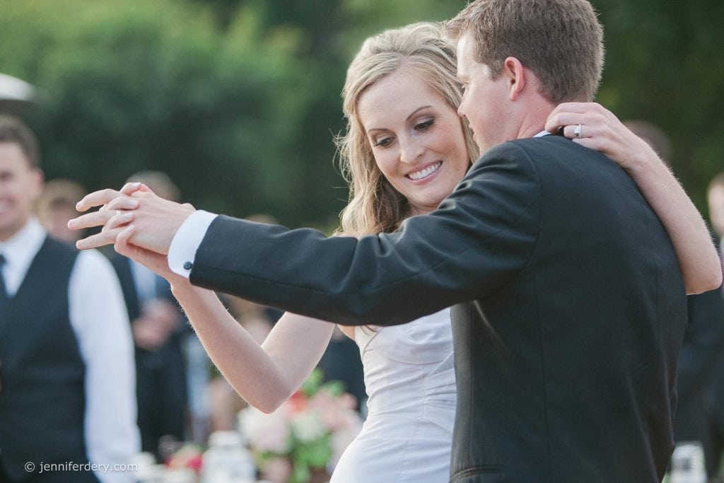 A bride and groom smile and dance closely together outdoors, dressed in a white wedding gown and a black suit, surrounded by blurred guests in the background.