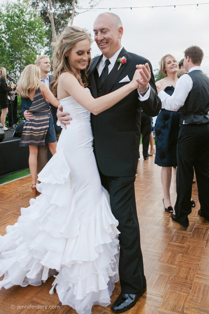 A smiling bride in a white ruffled gown dances with her father in a black suit on a wooden floor, surrounded by other guests at an outdoor wedding reception at Rancho Valencia Resort.