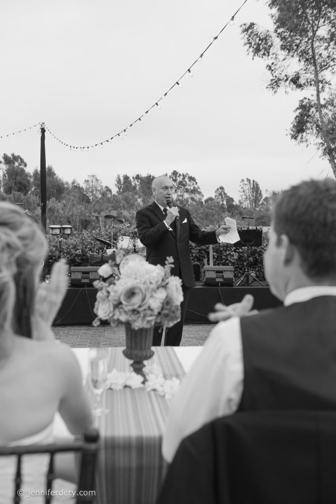 A man in a suit gives a speech outdoors at a wedding reception, holding a microphone and a piece of paper. A flower arrangement and two people facing him are visible in the foreground. String lights hang overhead.