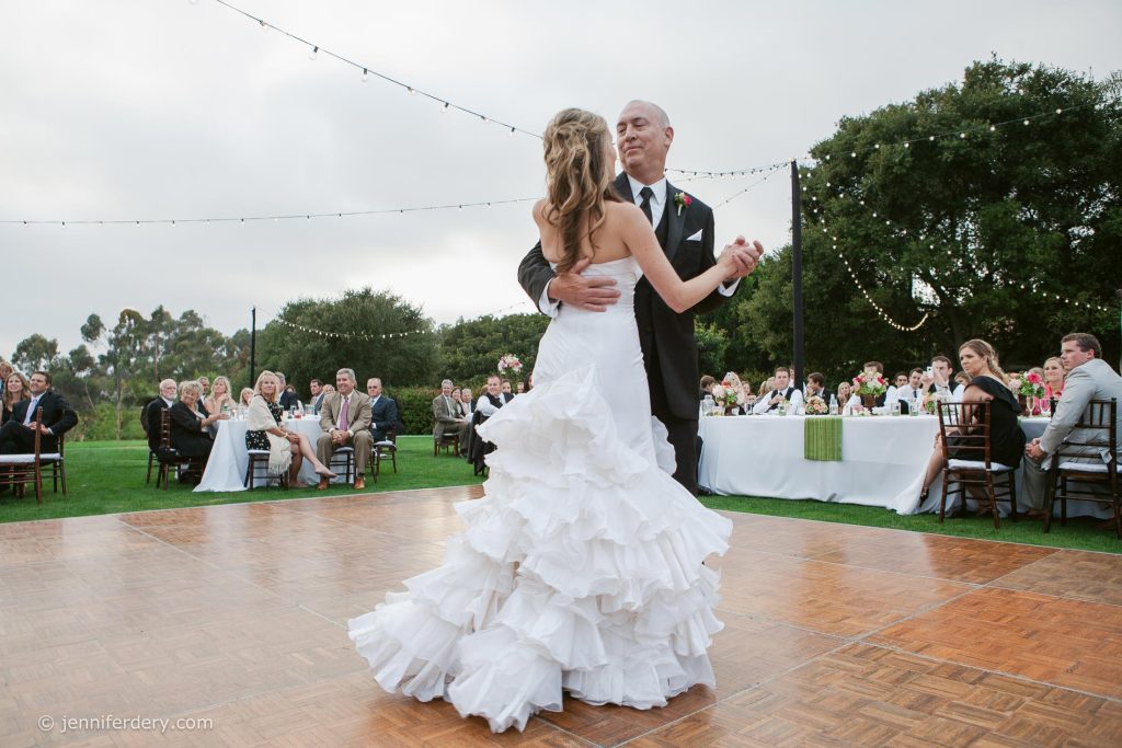 A bride in a ruffled white wedding dress dances with her father in a suit on an outdoor dance floor at Rancho Valencia Resort, surrounded by seated guests and string lights at a wedding reception.
