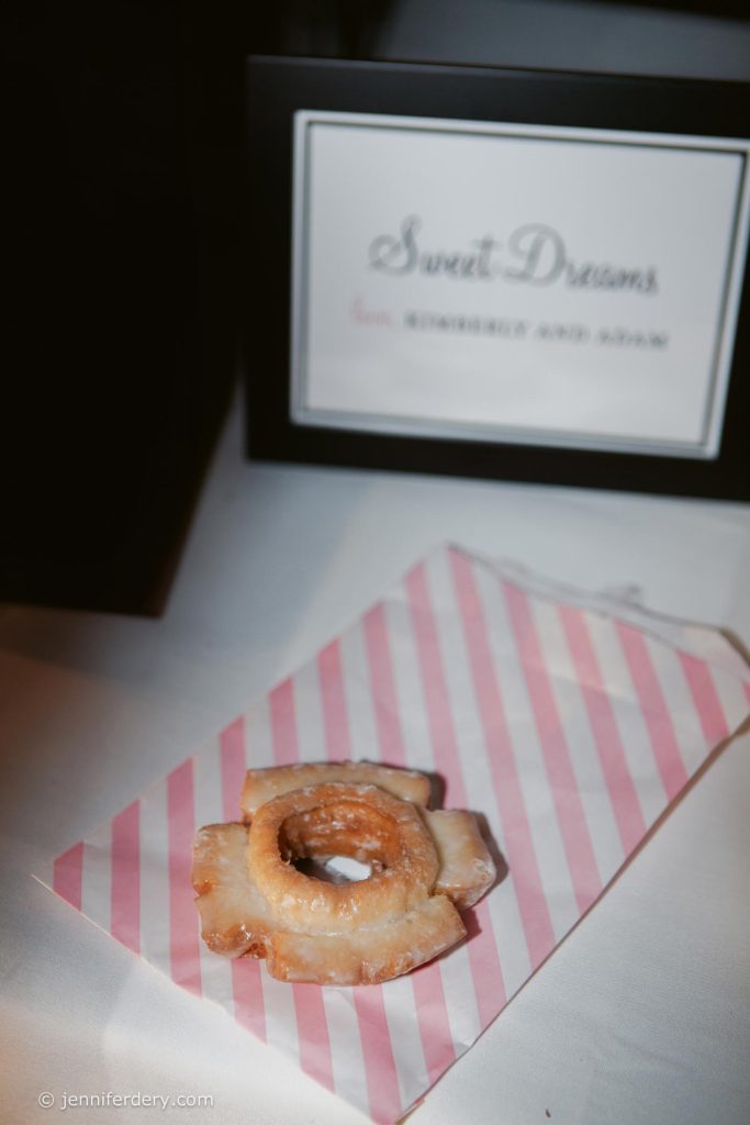 A glazed donut sits on a pink and white striped paper bag, with a framed sign in the background that reads "Sweet Dreams" and partially visible names.