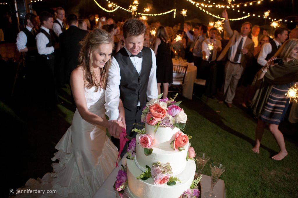 A bride and groom smile as they cut a tiered wedding cake decorated with flowers at an outdoor nighttime reception, surrounded by guests holding sparklers under string lights.