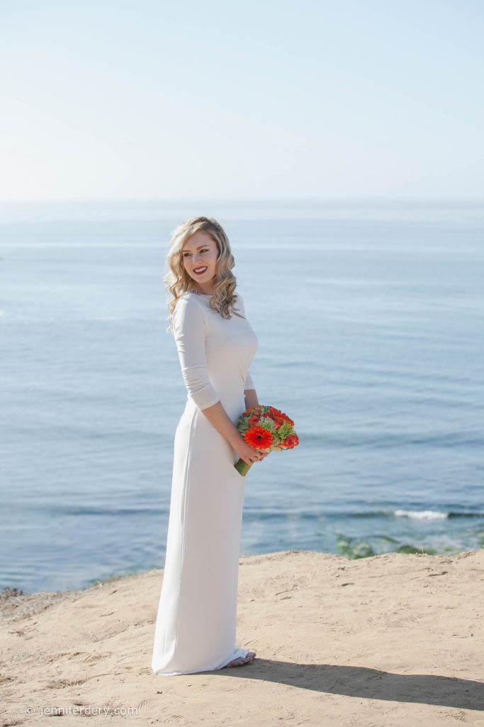 A woman in a long white dress stands on a sandy cliff overlooking a calm ocean, holding a bouquet of orange flowers and smiling at the camera under a clear blue sky.