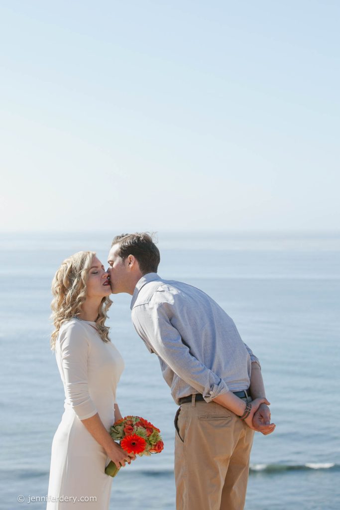 A man kisses a woman on the cheek as she smiles and holds an orange bouquet. They stand outdoors with the ocean and clear sky in the background.