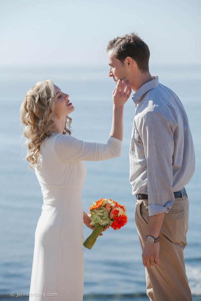 A bride in a white dress holds a bouquet of orange flowers and gently touches the groom's chin as they smile at each other by the water on a sunny day.