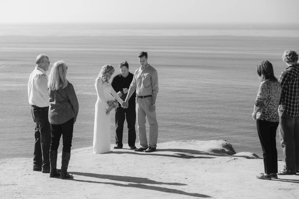 A small wedding ceremony takes place on a cliff overlooking the ocean. The couple stands holding hands with an officiant, while four guests stand nearby, all facing the couple. The scene is in black and white.