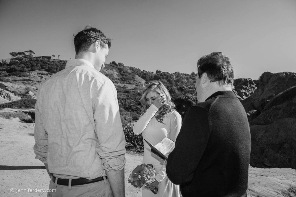 A woman wipes away a tear while holding a bouquet, standing outdoors between two men, one reading from a paper. All are dressed semi-formally against a backdrop of cliffs and trees under a clear sky.