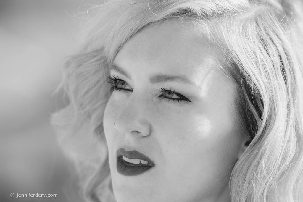 Black and white close-up of a woman with wavy, light-colored hair, dark lipstick, and defined eyelashes, gazing into the distance with a thoughtful expression. © jenniferdery.com appears in the lower left corner.