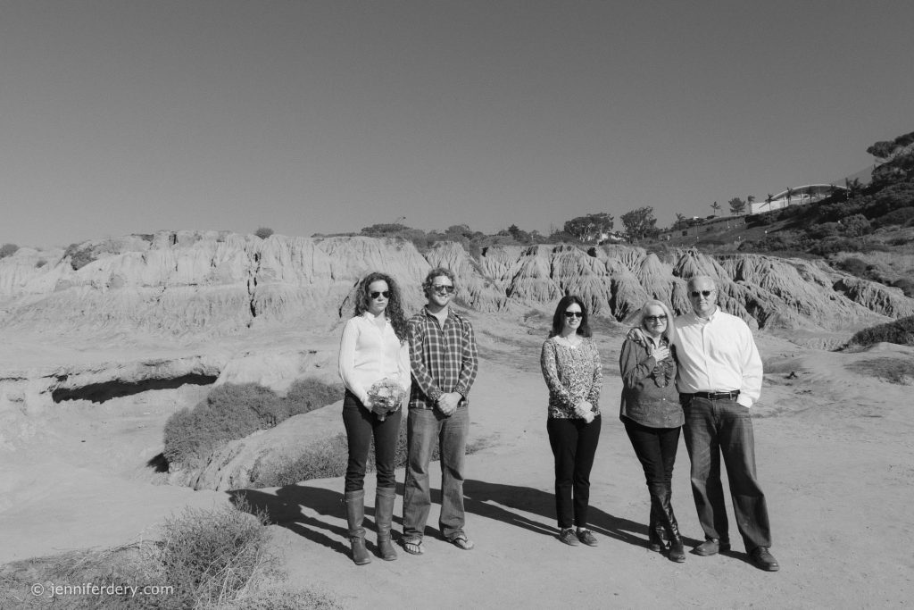 Five people stand together outdoors in a rocky, desert-like landscape under a clear sky. They wear casual clothing and sunglasses, facing the camera with neutral expressions. Shrubs and cliffs are in the background.