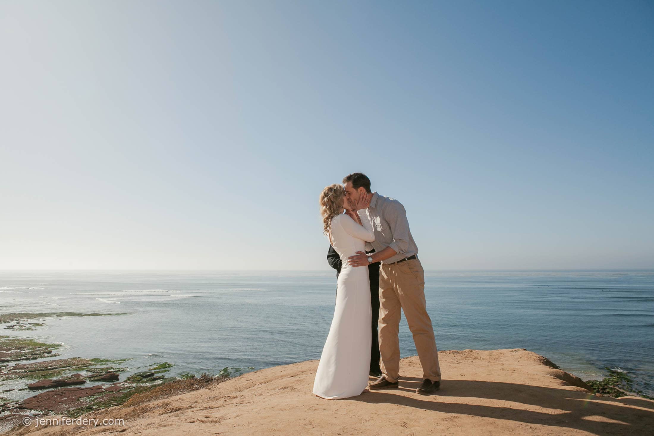 A couple stands at the edge of a cliff by the ocean, embracing and kissing under a clear blue sky, with the sea and shoreline visible in the background.