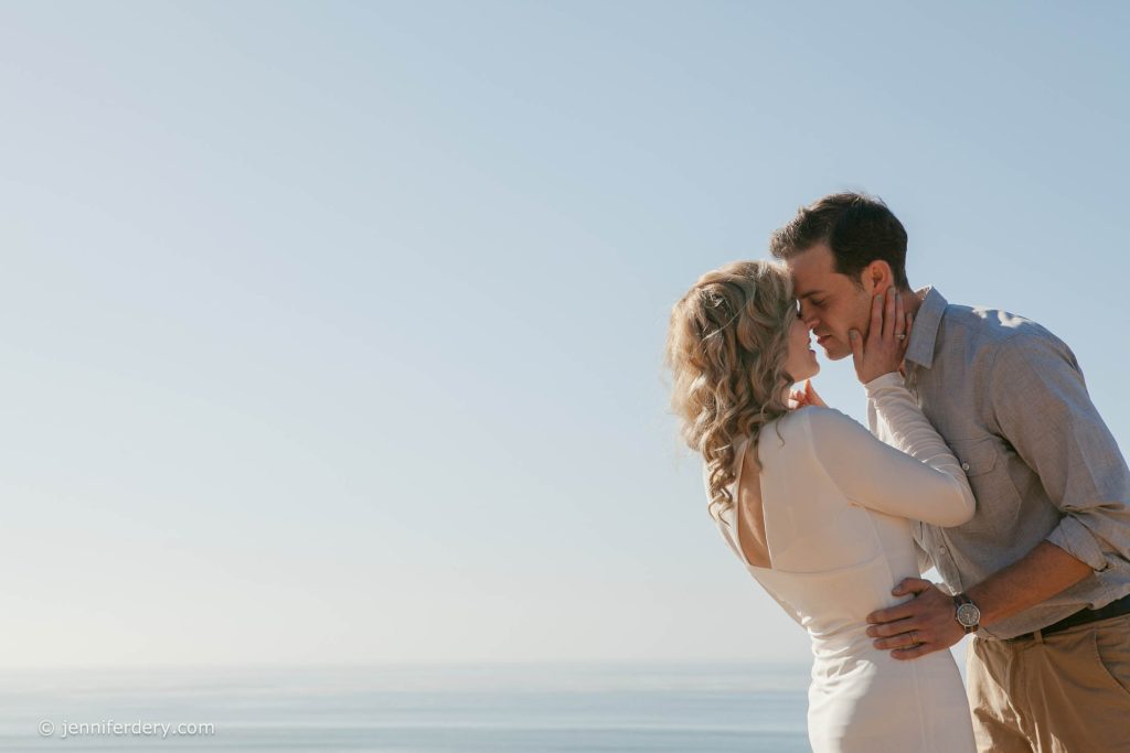 A couple embraces and kisses by the ocean under a clear blue sky, with the woman in a white dress and the man in a light shirt and khakis.