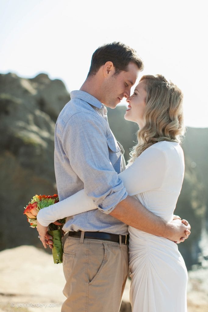 A couple embraces outdoors on a sunny day, standing close and smiling with foreheads touching. The woman in a white dress holds a small bouquet of flowers behind the man's back. Rocky cliffs are visible in the background.