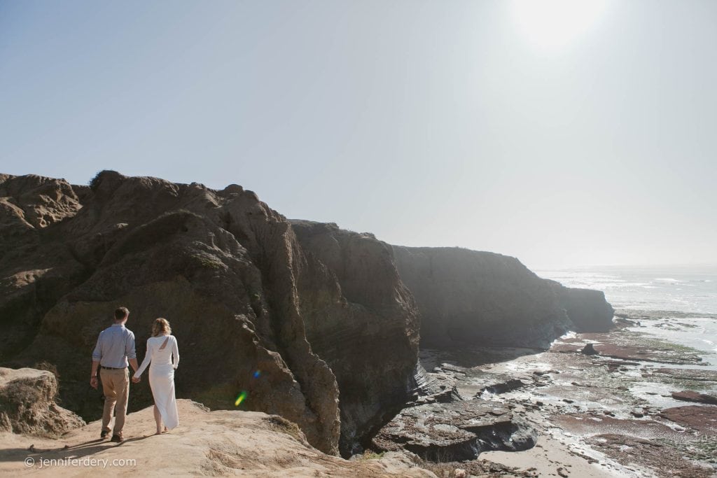 A couple holding hands walks along a rocky cliff overlooking the ocean, with sunlight shining brightly in the sky and tidal pools visible below.