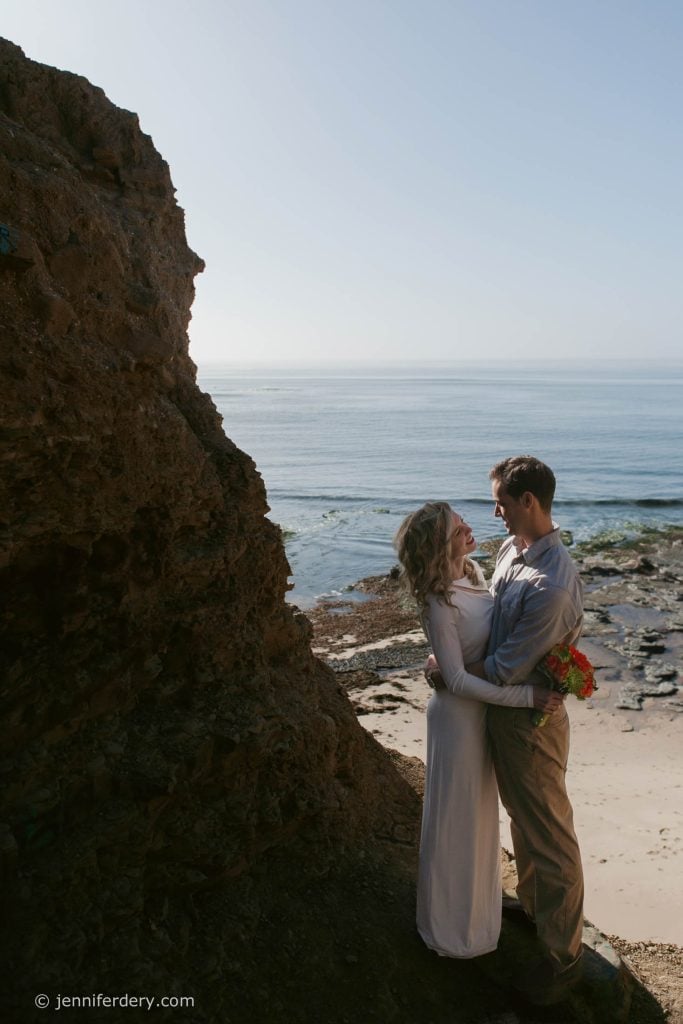 A couple embraces and smiles at each other on a rocky beach, with the ocean and blue sky in the background. The woman holds a bouquet of flowers. Sunlight creates a serene atmosphere.