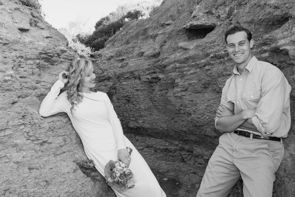 A woman in a long white dress holding a bouquet sits on a rocky ledge, smiling at a man in a button-down shirt and pants who stands nearby with arms crossed, both outdoors in a rocky setting.
