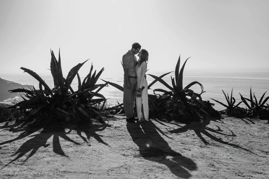 A couple stands close together, touching foreheads, between large plants on a cliff overlooking the ocean. Their shadows stretch toward the camera in the sunlight. The image is in black and white.