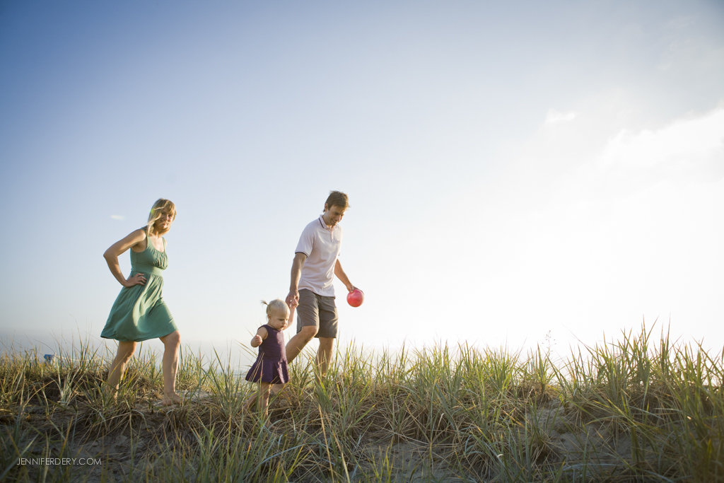 A family walking on a grassy sand dune. A woman in a green dress, a man in a white shirt holding a red frisbee, and a toddler in a purple dress are enjoying a sunny day under a clear blue sky.