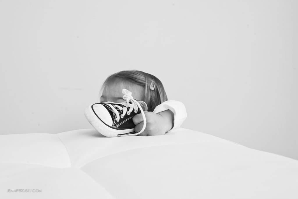 A small child peeks over a cushioned surface, holding a black and white sneaker with a playful expression. The background is plain, emphasizing the child's curiosity and the shoe. Black and white photo.