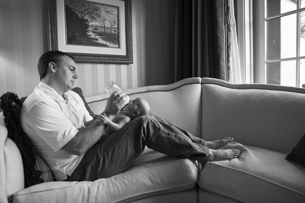 A man sits on a couch feeding a baby from a bottle. They are in a cozy room with a painting on the wall and sunlight streaming through a window.