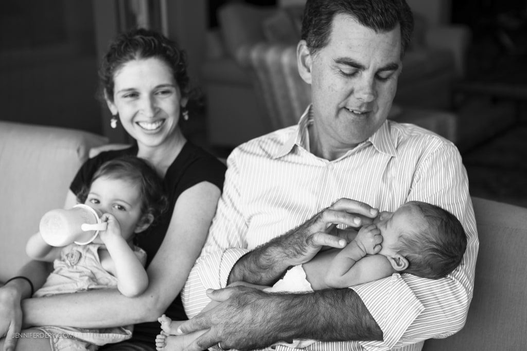 A smiling woman sits beside a man holding a newborn. The woman holds a toddler drinking from a bottle on her lap. They are seated on a couch in a warmly lit room.