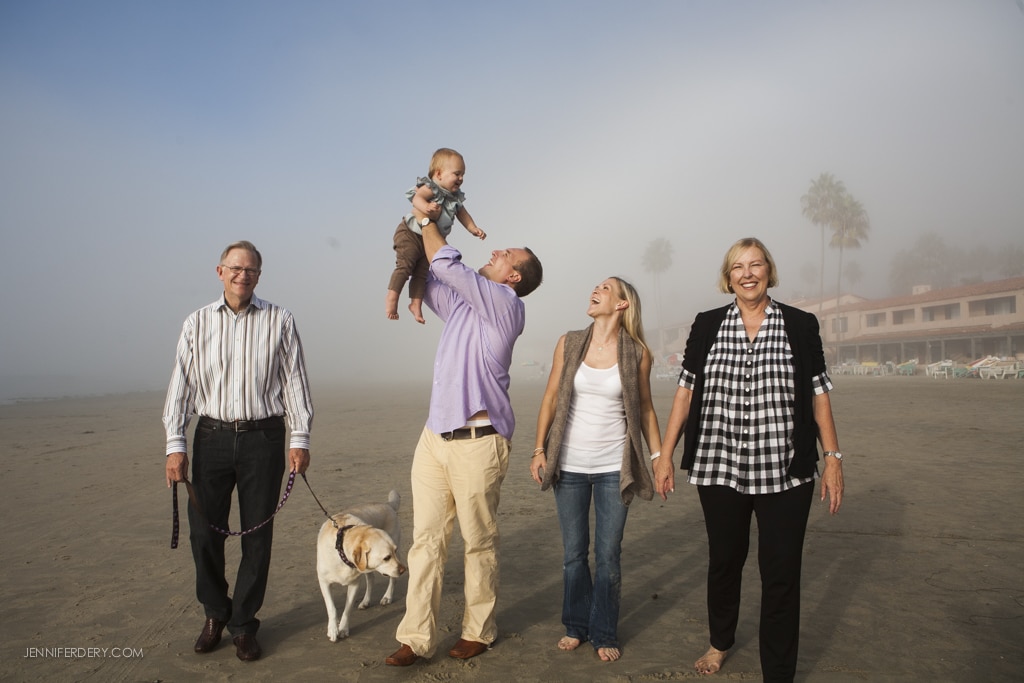 A family enjoys a day at the beach. A man lifts a baby while a woman smiles at them. Two other adults walk beside them, one holding a dog on a leash. The background shows misty beach scenery with palm trees and buildings.