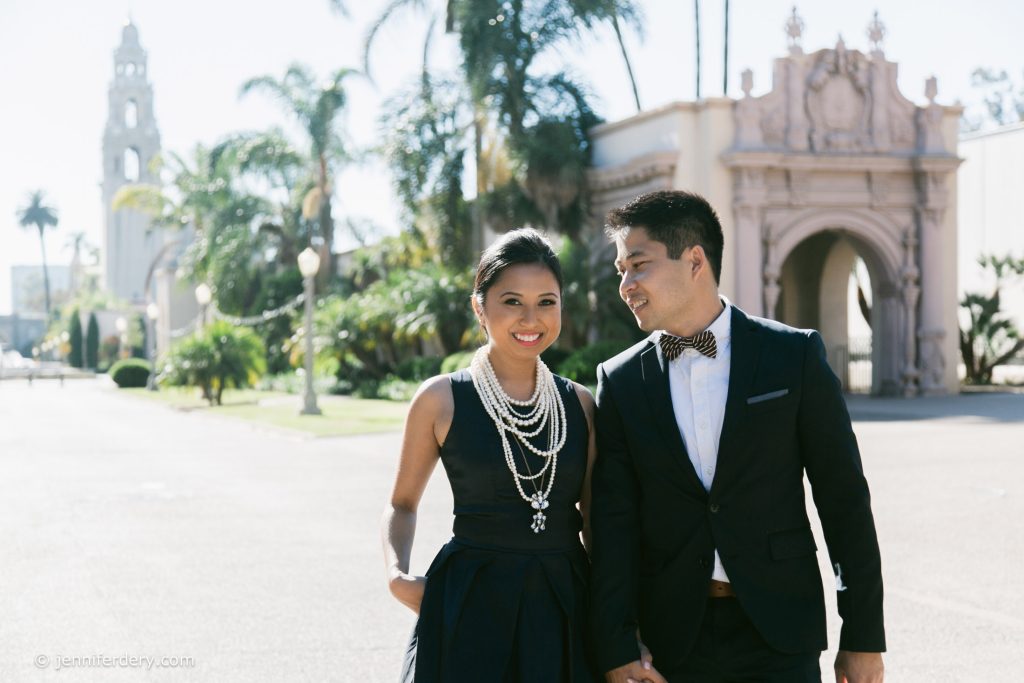 A well-dressed couple walks outdoors on a sunny day, smiling. The woman wears a dark dress with layered pearl necklaces; the man wears a dark suit and bow tie. Palm trees and an ornate building are visible in the background.