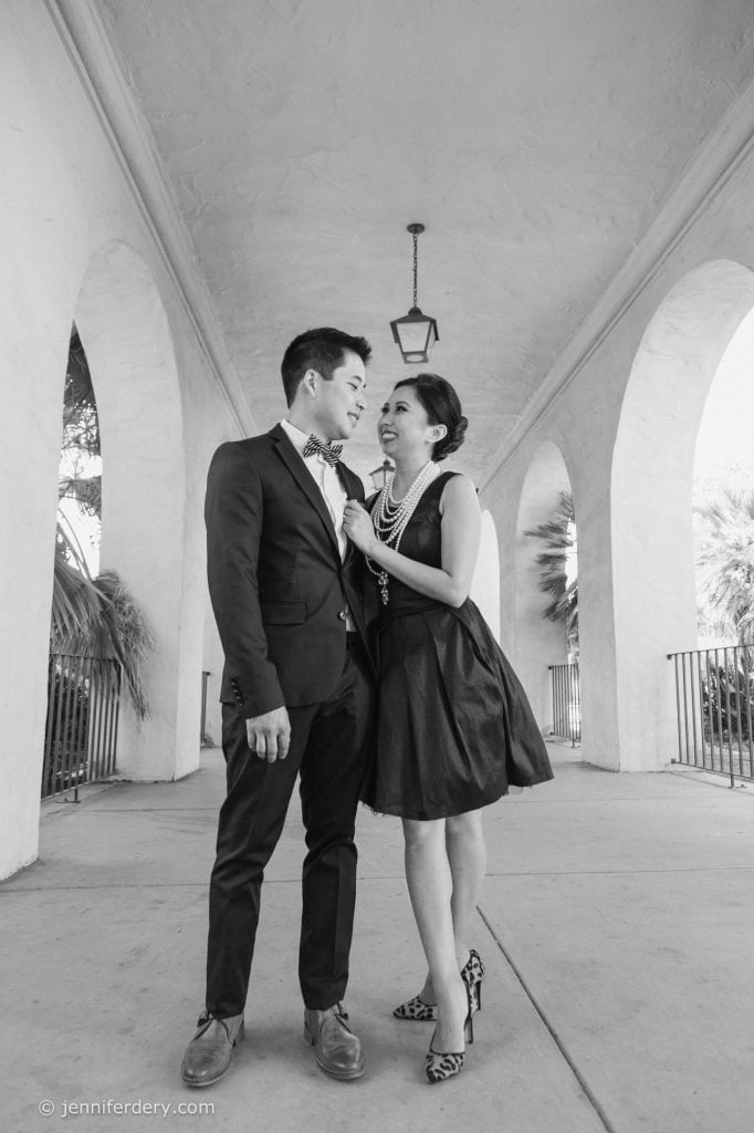 A couple dressed formally stand under an arched walkway, smiling at each other. The woman touches the man's bow tie, and both appear happy and affectionate. The photo is in black and white.