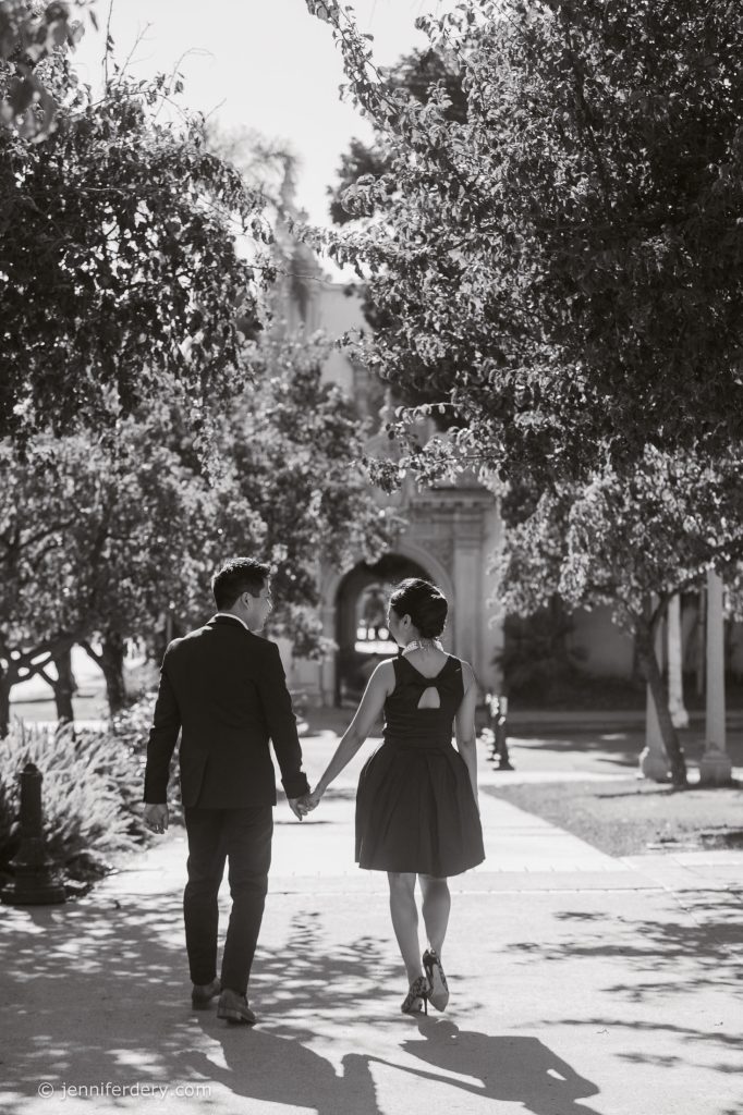 A couple dressed formally walks hand-in-hand down a sunlit path lined with trees, their backs to the camera. Shadows and dappled sunlight create a romantic atmosphere in this black-and-white photo.