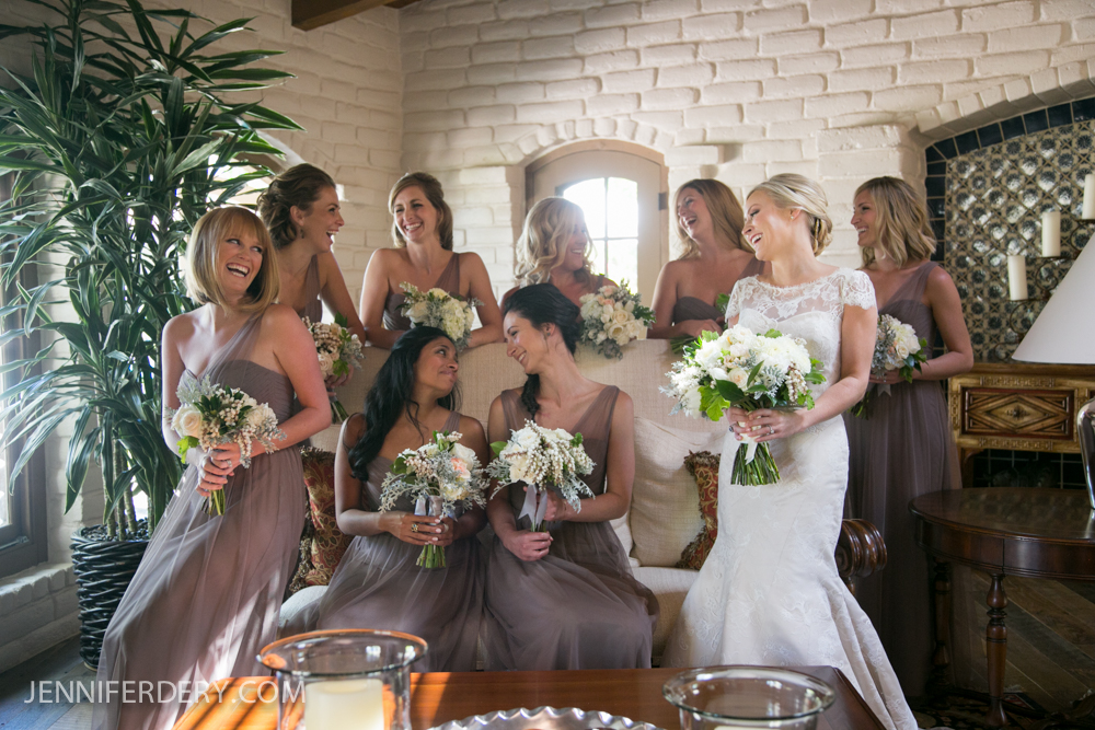 A bride in a white dress stands smiling with her bridesmaids, who are wearing matching taupe dresses and holding bouquets, gathered together in a bright, warmly lit room with white brick walls.