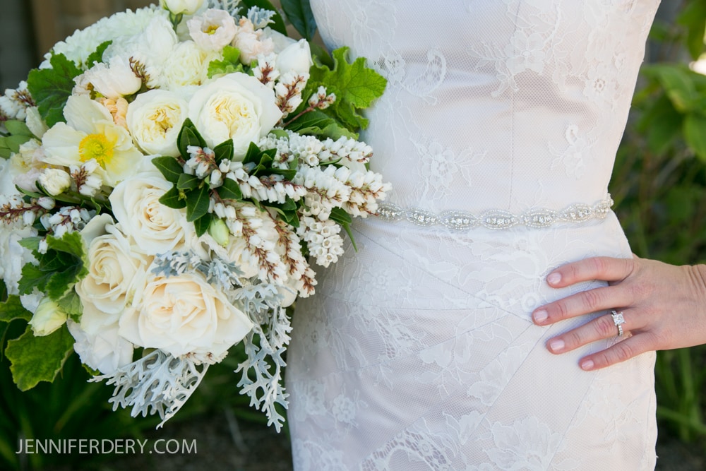 A bride in a lace wedding dress holds a bouquet of white and cream flowers with greenery. Her left hand, showing a diamond ring, rests on her hip. The text “JENNIFERDERY.COM” appears at the bottom left.