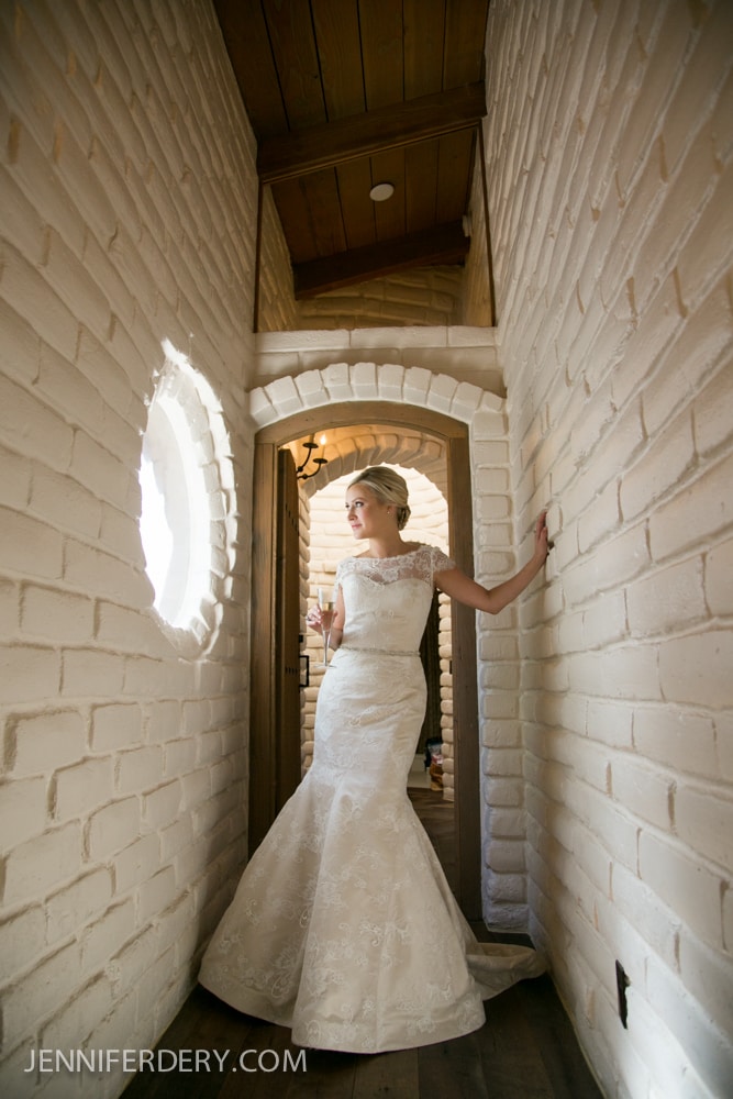 A bride in a white lace gown stands in a narrow hallway with white brick walls, softly lit by sunlight from a round window, gazing thoughtfully to the side, holding a glass.