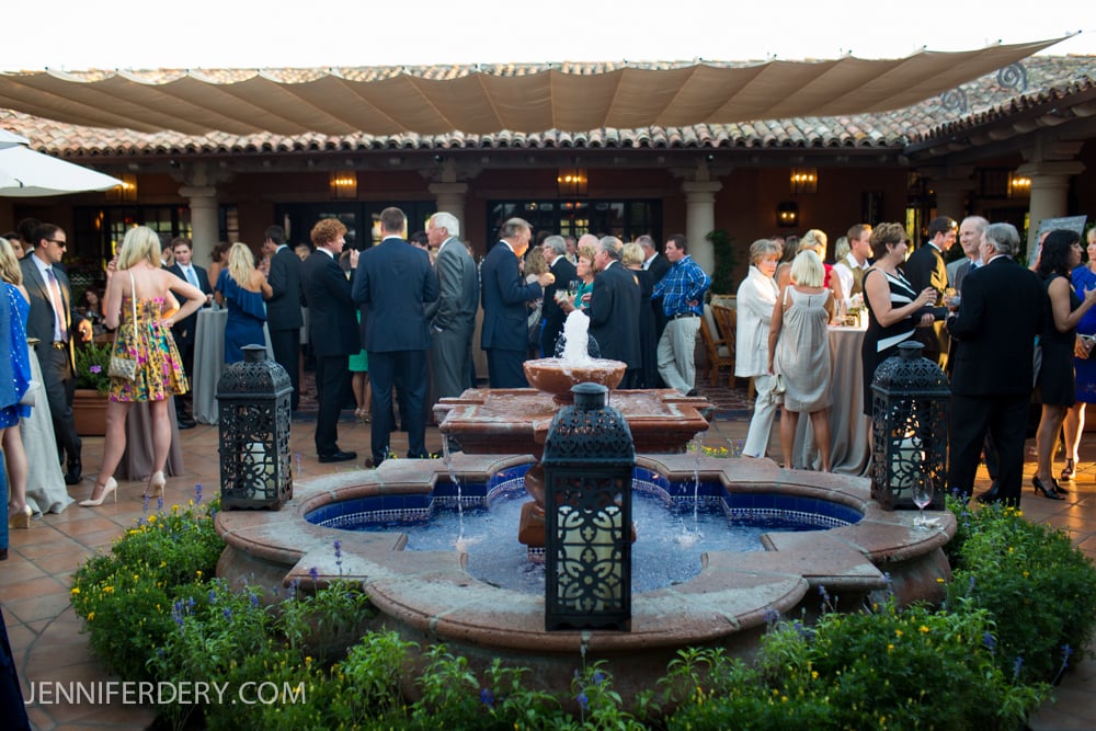 A group of people in formal attire socialize around a large stone fountain at an outdoor event with a tiled roof patio and covered terrace in the background.