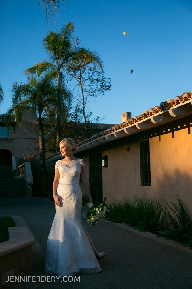 A bride in a white lace gown stands outside a sunlit building with a tile roof, holding a bouquet. Palm trees and hot air balloons are visible in the clear blue sky.