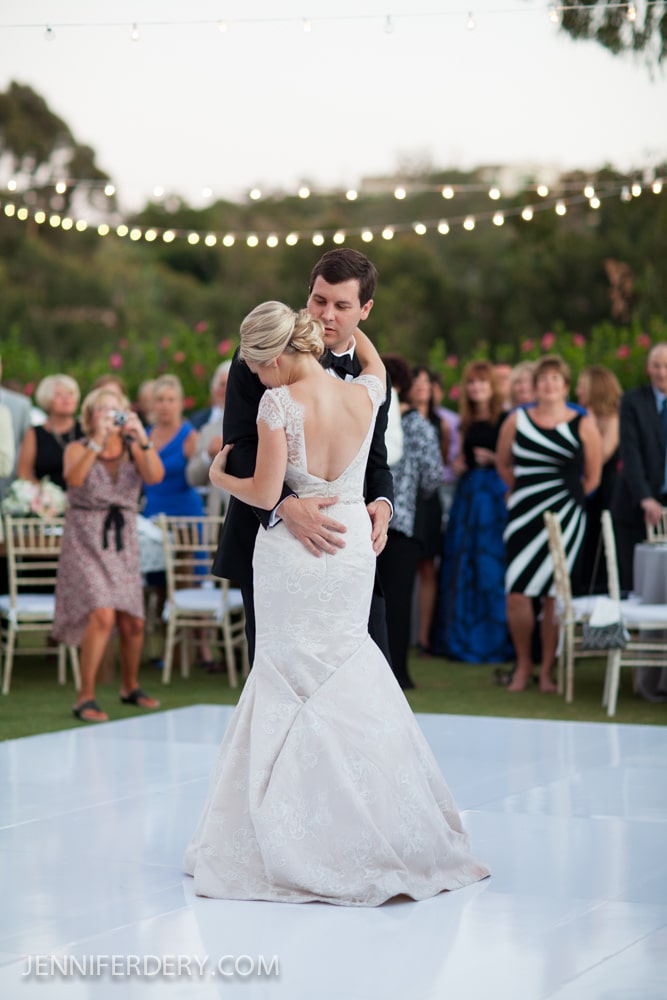 A bride and groom share their first dance outdoors at their wedding on the Rancho Valencia croquet Lawn, surrounded by seated and standing guests. String lights are overhead, and the bride wears a fitted, open-back gown. The groom wears a black tuxedo.