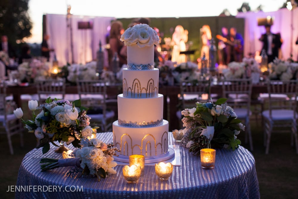 A three-tiered white wedding cake with blue and gold accents sits on a table decorated with candles and floral arrangements, set outdoors with blurred guests and a band in the background.