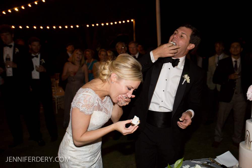 A bride and groom laugh as they feed each other cake at their nighttime wedding reception, surrounded by guests. The bride wears a white lace dress; the groom wears a tuxedo.