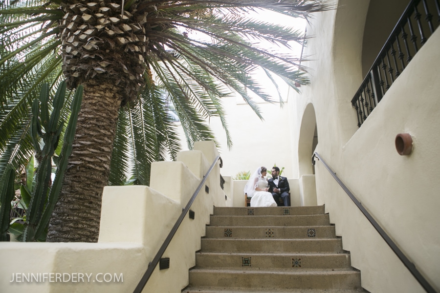 A bride and groom sit together on outdoor stairs beneath a large palm tree at their Estancia La Jolla Wedding, surrounded by cream-colored walls and greenery, sharing a quiet moment on their special day.