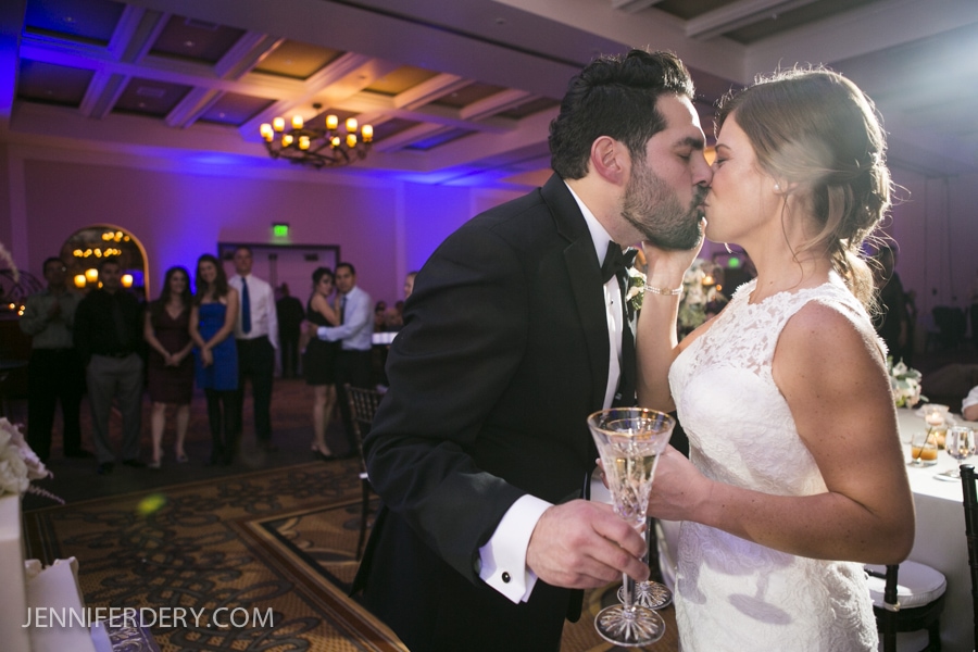 A bride and groom in formal wedding attire share a kiss while holding champagne glasses at their Estancia La Jolla Wedding reception, with guests and elegant decor visible in the background.
