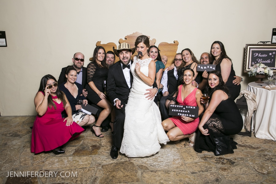 A bride and groom pose playfully with a large group of friends at their Estancia La Jolla Wedding, some holding funny signs. Everyone is smiling in formal attire, gathered indoors against a light wall for this joyful celebration.