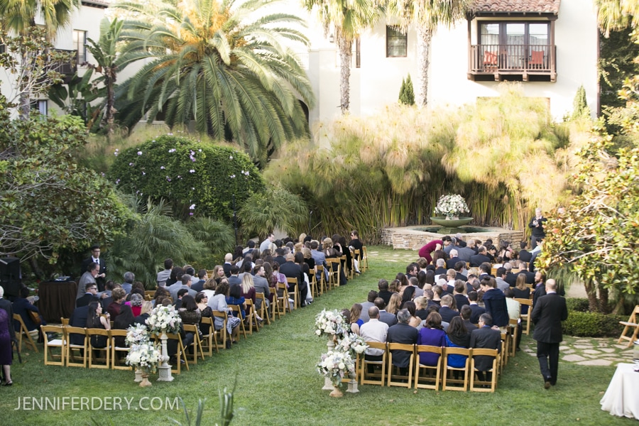 An outdoor wedding ceremony at Estancia La Jolla with guests seated in rows of wooden chairs on a lawn, surrounded by lush greenery and palm trees. A flower arrangement is at the front, and a building is visible in the background.