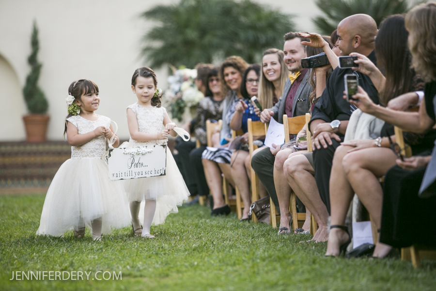 Two young flower girls in white dresses walk down an outdoor aisle, smiling, while guests seated on either side watch and take photos. One girl holds a white sign with text. It is a sunny wedding ceremony.