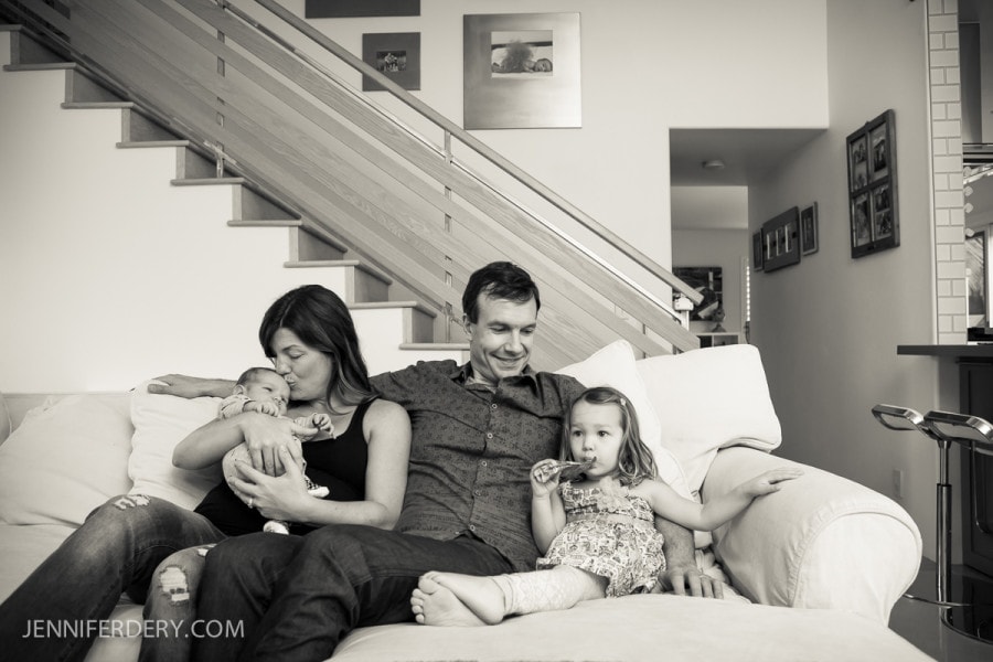 A family sits on a couch in a living room. The mother holds a baby, while the father sits next to her. A young girl sits beside the father, holding an object in her mouth. Stairs and framed pictures are visible in the background.