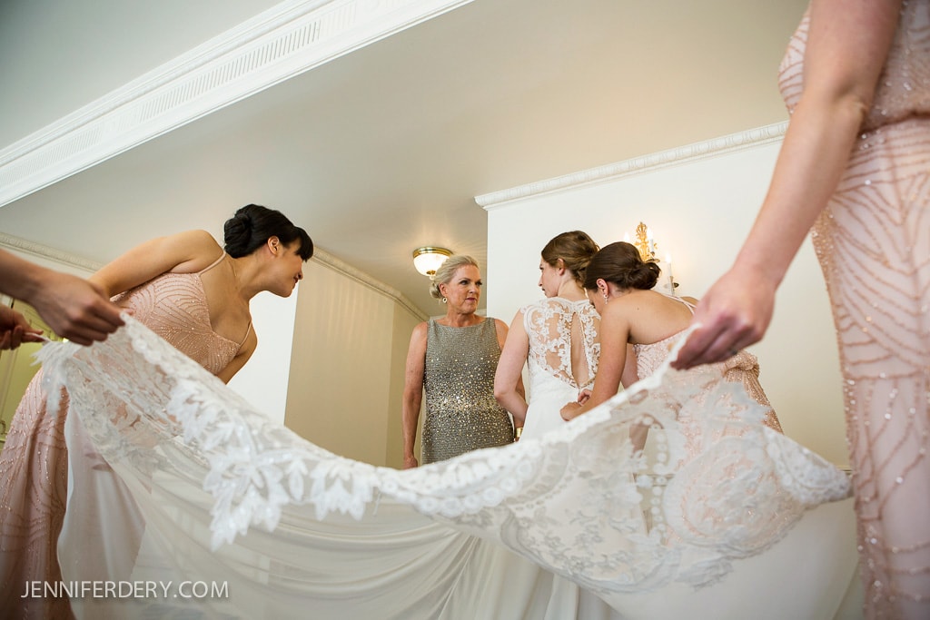 A bride dressed in white is assisted by four women wearing pastel dresses, who are adjusting her long, intricate lace veil. They are in a well-lit room with a white ceiling and light walls. A woman in a silver dress stands nearby, observing.